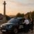 Professional Waltham Cross taxi driver opens the door for a business passenger in front of the Eleanor Cross monument, with futuristic AI dispatch and fixed fare holograms floating above the vehicle in a sunlit Hertfordshire street.